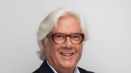 Mann mit grauen Haaren und Brille im dunkelblauen Sakko, lächelnd mit verschränkten Armen, Studio-Porträt vor hellem Hintergrund.