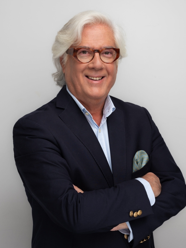 Mann mit grauen Haaren und Brille im dunkelblauen Sakko, l&auml;chelnd mit verschr&auml;nkten Armen, Studio-Portr&auml;t vor hellem Hintergrund.