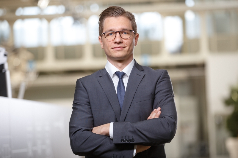Mann im Anzug mit Brille, Arme verschr&auml;nkt, steht in einem modernen B&uuml;ro mit unscharfem Hintergrund.