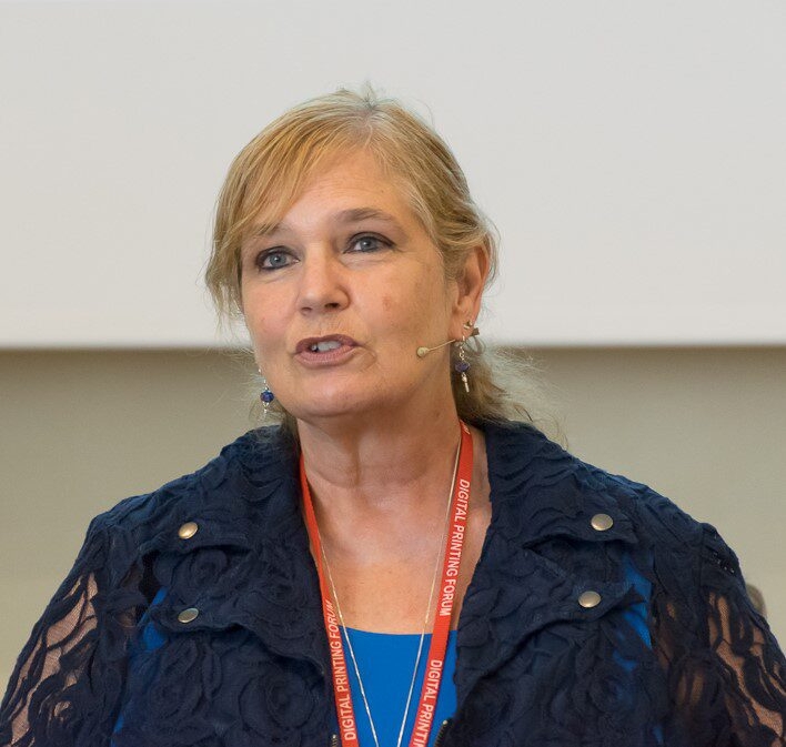 Frau mit Headset-Mikrofon spricht bei einer Veranstaltung; tr&auml;gt dunkelblaue Jacke und orangefarbenes Lanyard.